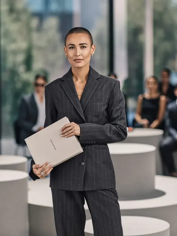Une femme en tailleur gris à fines rayures tient un dossier devant des personnes assises, floues, à l'arrière-plan